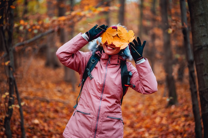 Beautiful happy blond adult woman with a smile holds autumn yellow orange leaves hiding her face with fall colored forest trees in background