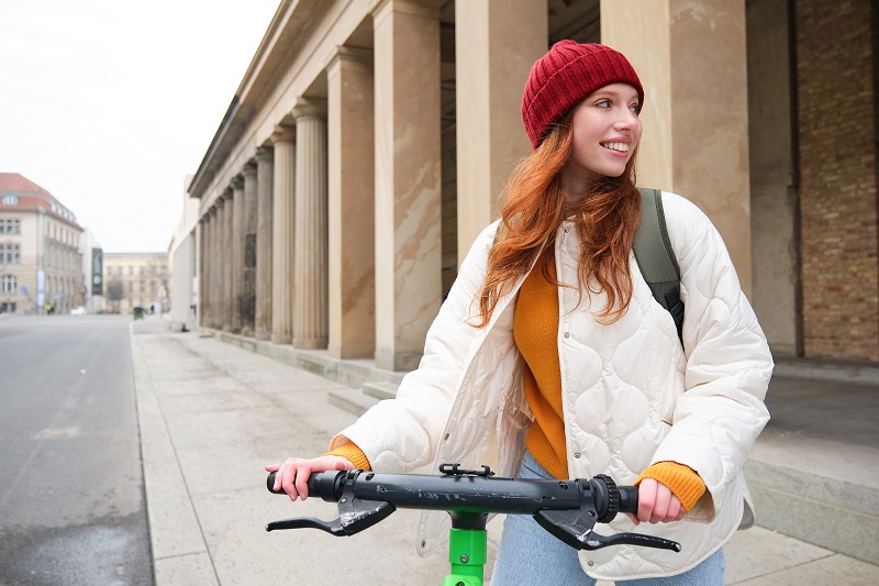 Young smiling redhead girl, student rides electric scooter, rents it and travels around city